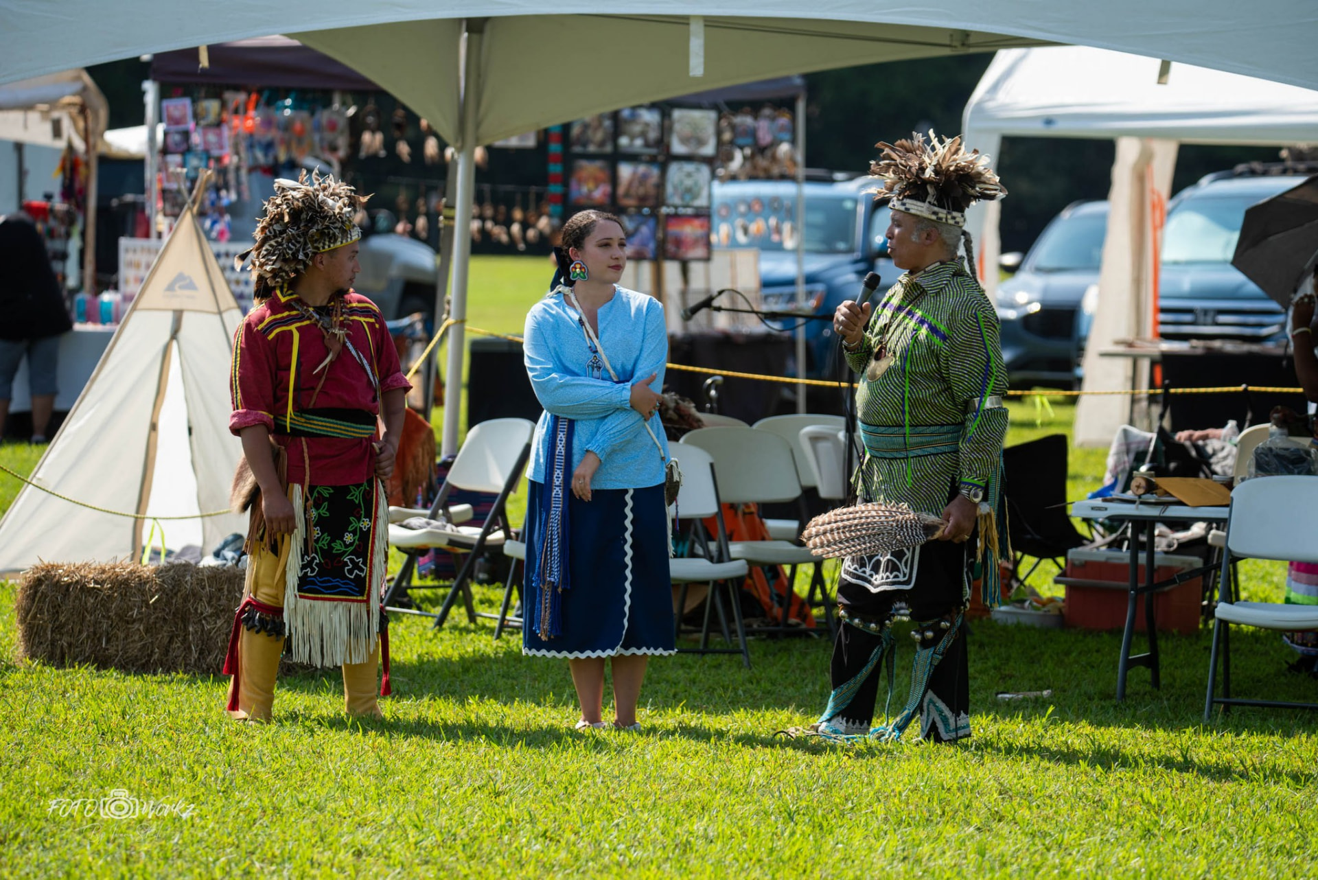 Three people standing in a line under a white tent at an outdoor powwow, two of them wearing feathered headdresses