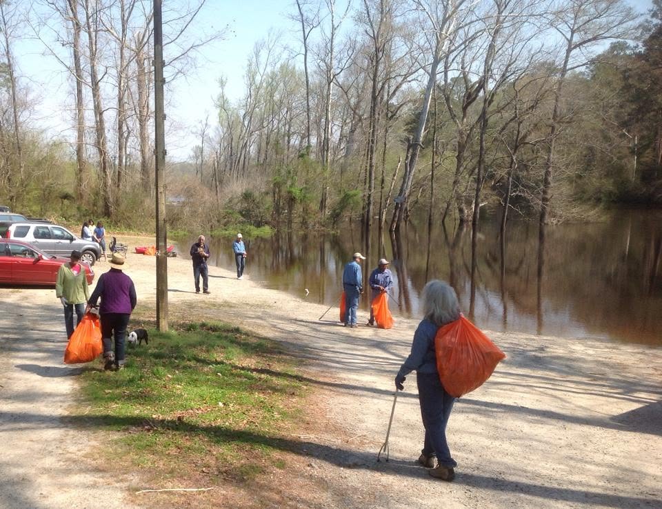 Volunteers standing on the gravel shore with filled orange trash bags, a small boat resting on the bank behind them