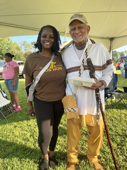 A young woman in a brown Nottoway Tribe of Virginia Pow Wow t-shirt and a smiling man in a tan Nottoway Indian Tribe cap stand together under a white tent