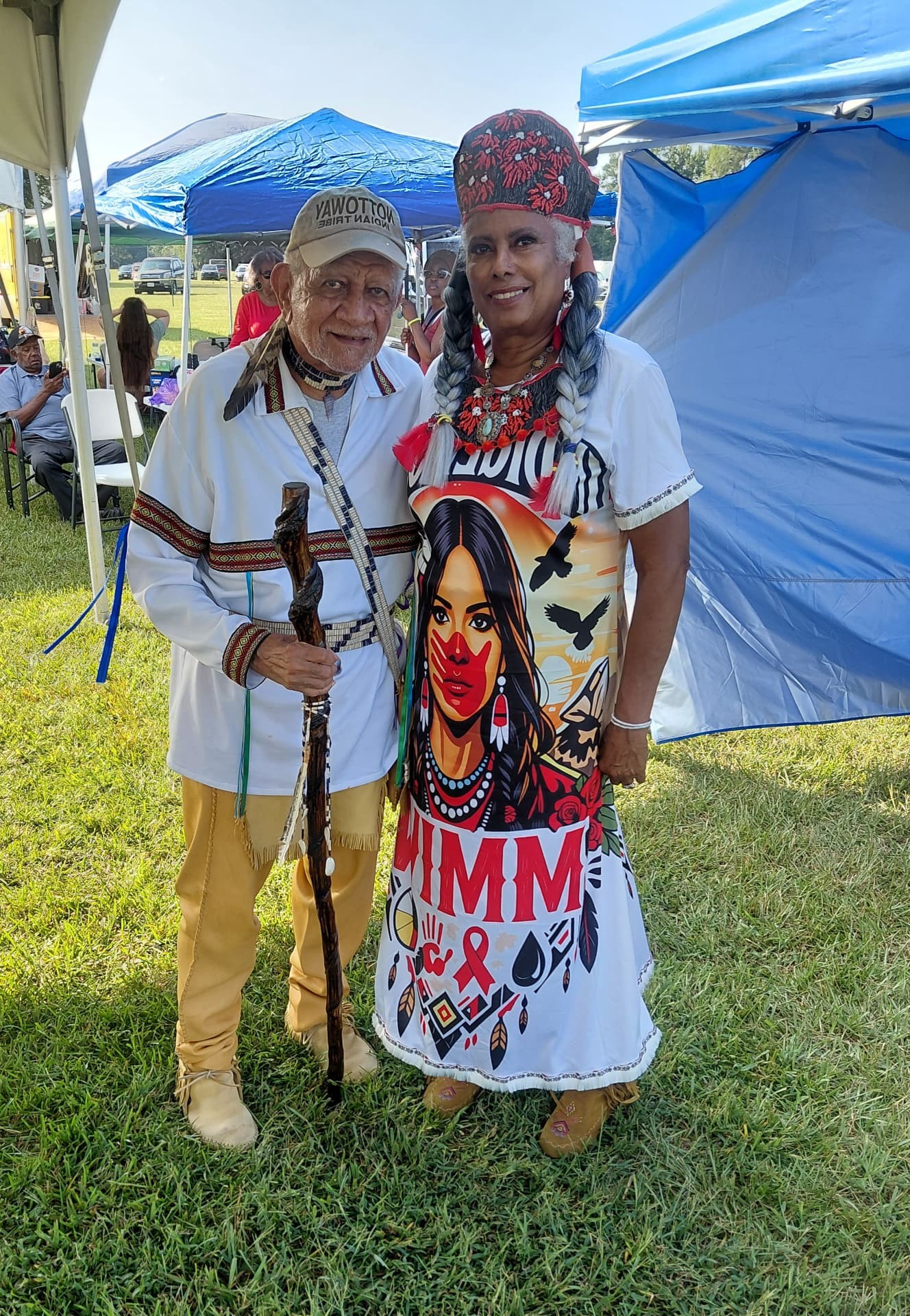 A tribal elder in a Nottoway cap holding his carved walking stick beside a tribal member in her beaded crown and regalia dress under the blue canopy
