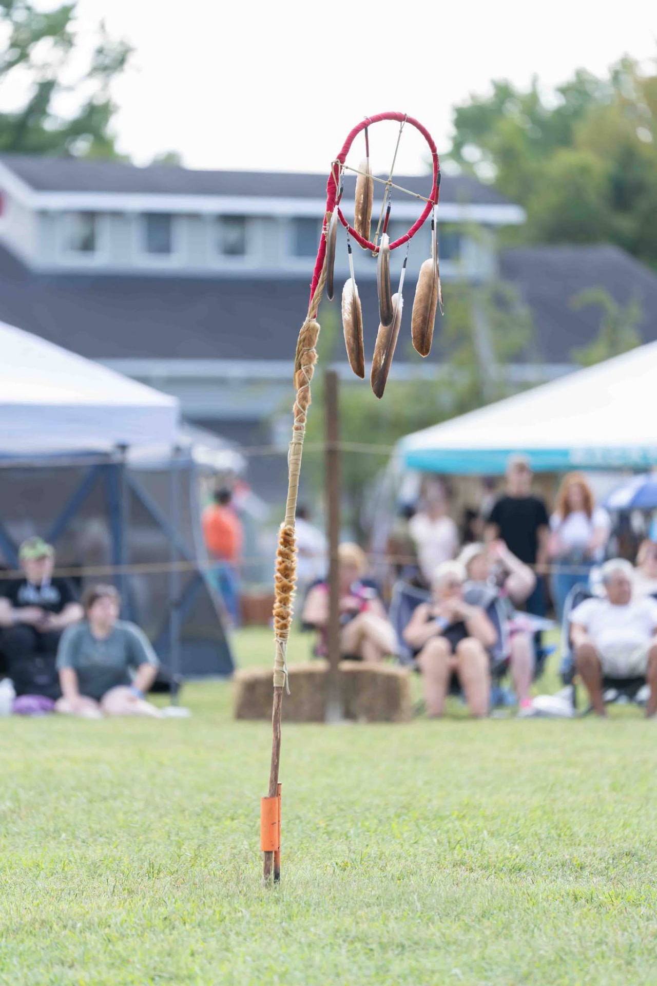 A dreamcatcher staff standing in the grass in front of the seated audience at the powwow