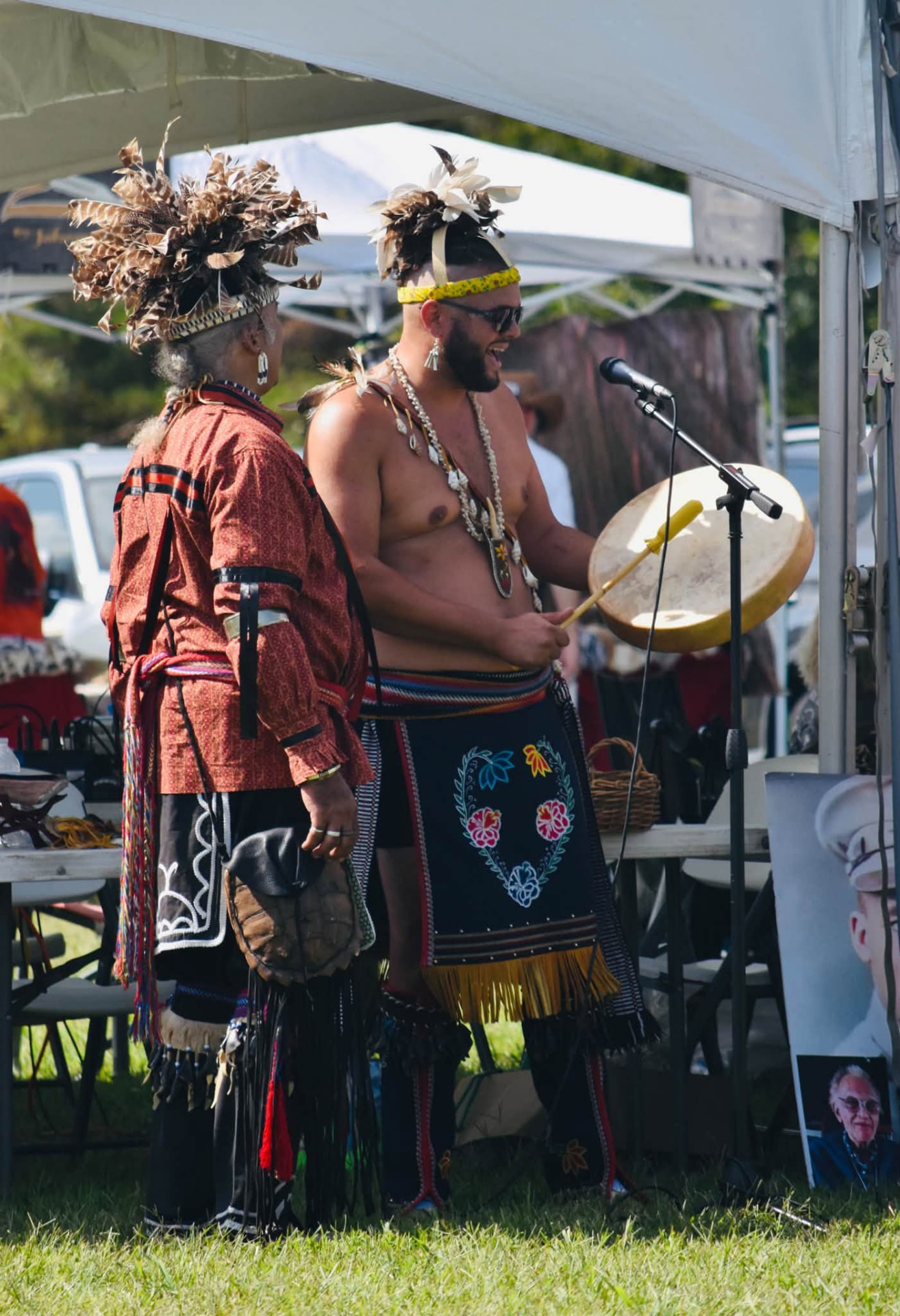 A tribal elder in a feathered headdress singing at the microphone beside a younger man holding a hand drum under the tent