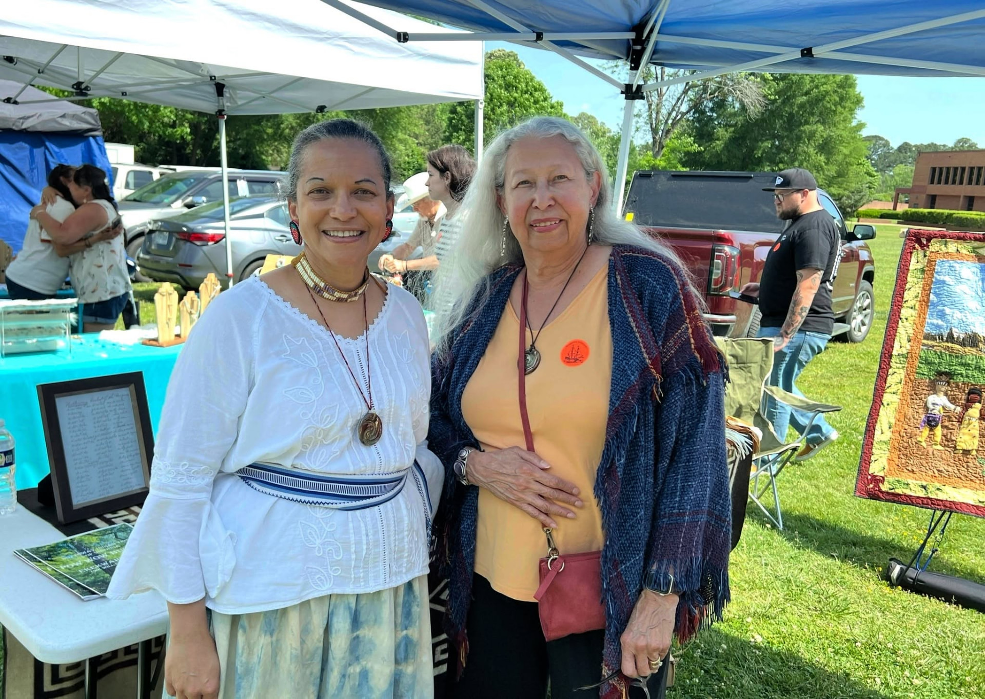 Two women stand behind the tribe's booth at the Jamestown Indigenous Arts Festival, with jewelry on stands and a woven wall hanging on display