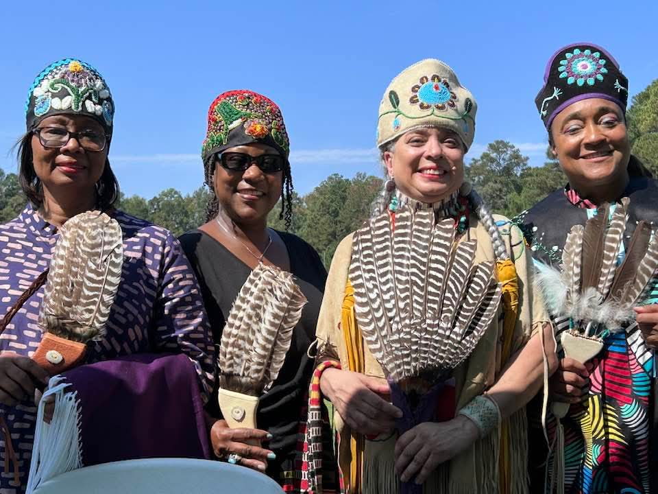 Four smiling women in a row, each wearing a beaded cap and holding a feather fan