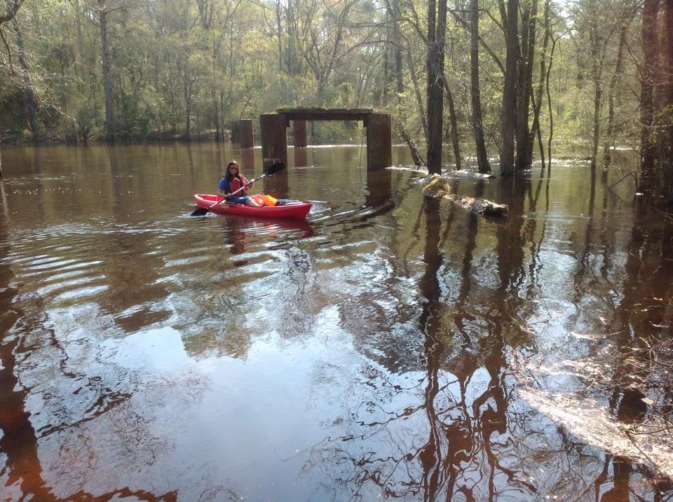 A volunteer in a red kayak on the Nottoway River below the metal pilings of Cary's Bridge, holding up a bag of litter