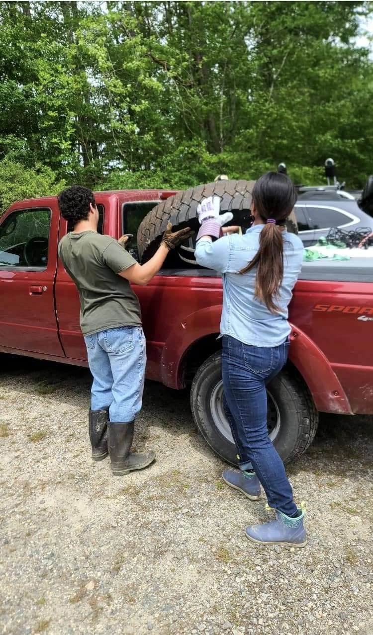 Two people lifting a mud-covered tire into the bed of a red pickup truck after the cleanup