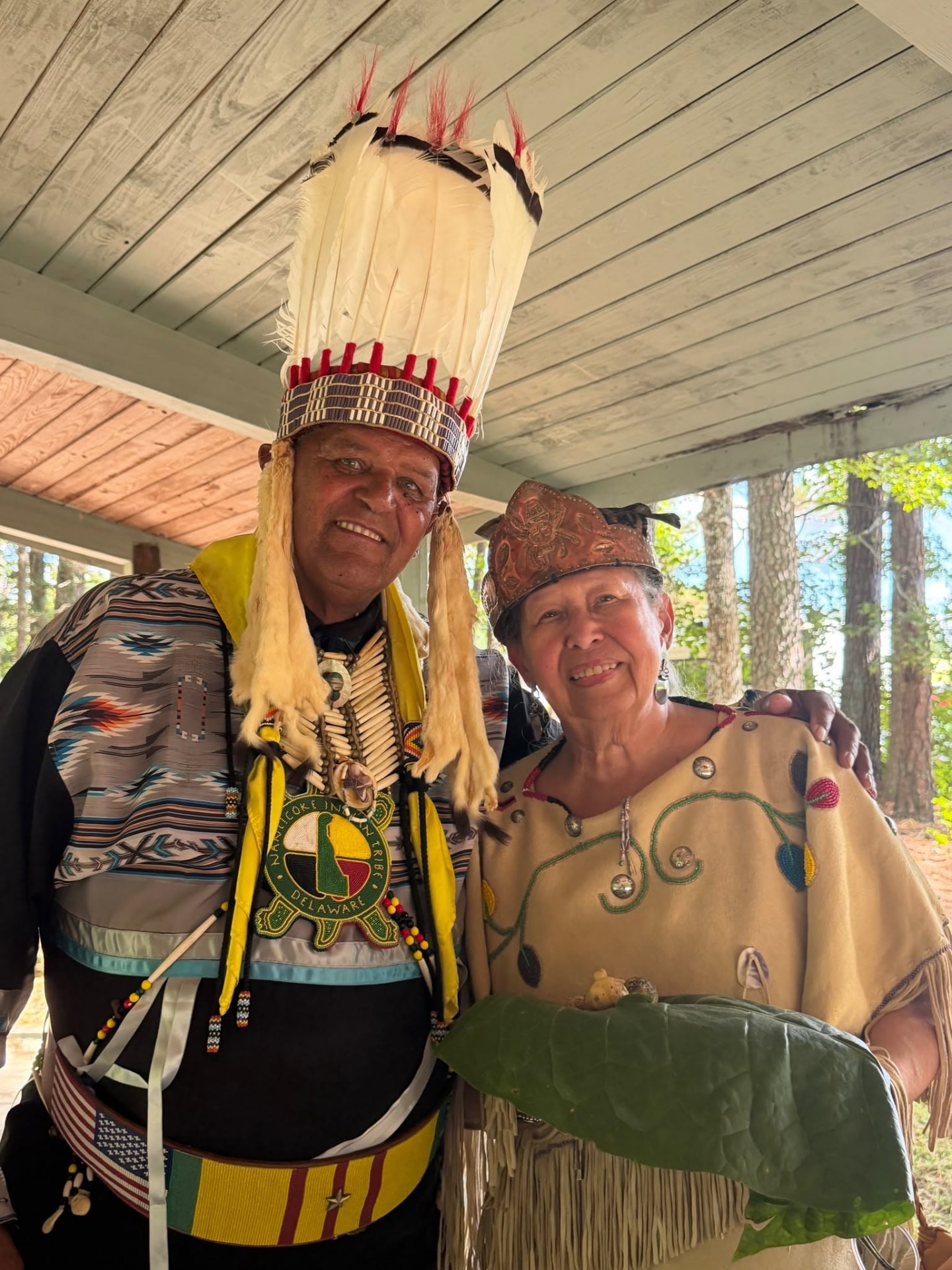 Two tribal elders smiling together — one in a feathered headdress and yellow shawl, the other in a beaded leather dress holding tobacco leaves
