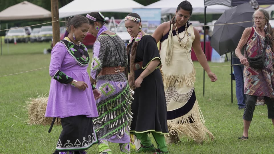 Four women in jingle dresses and beaded headbands dance in a row on a grassy field at a powwow