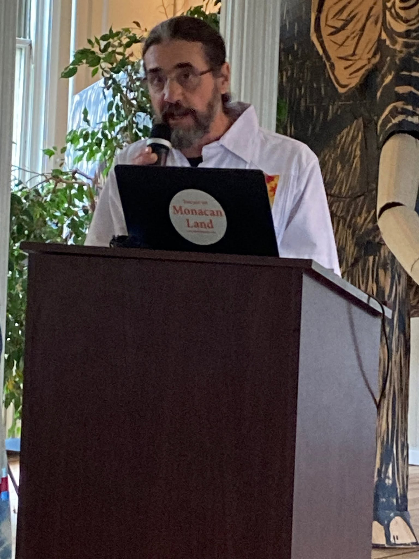 A man in glasses and a white shirt speaks into a microphone at a lectern during the Nottoway language presentation
