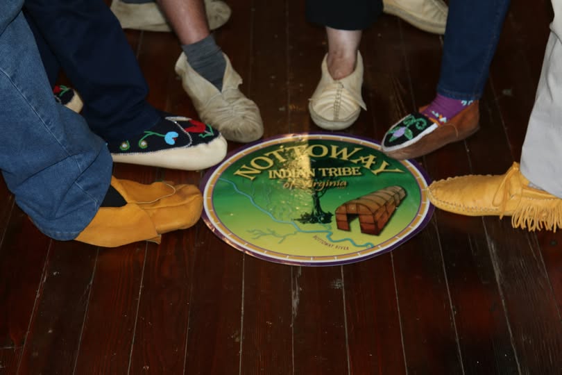 Five pairs of moccasins placed around the Nottoway Indian Tribe of Virginia seal on a polished wood floor