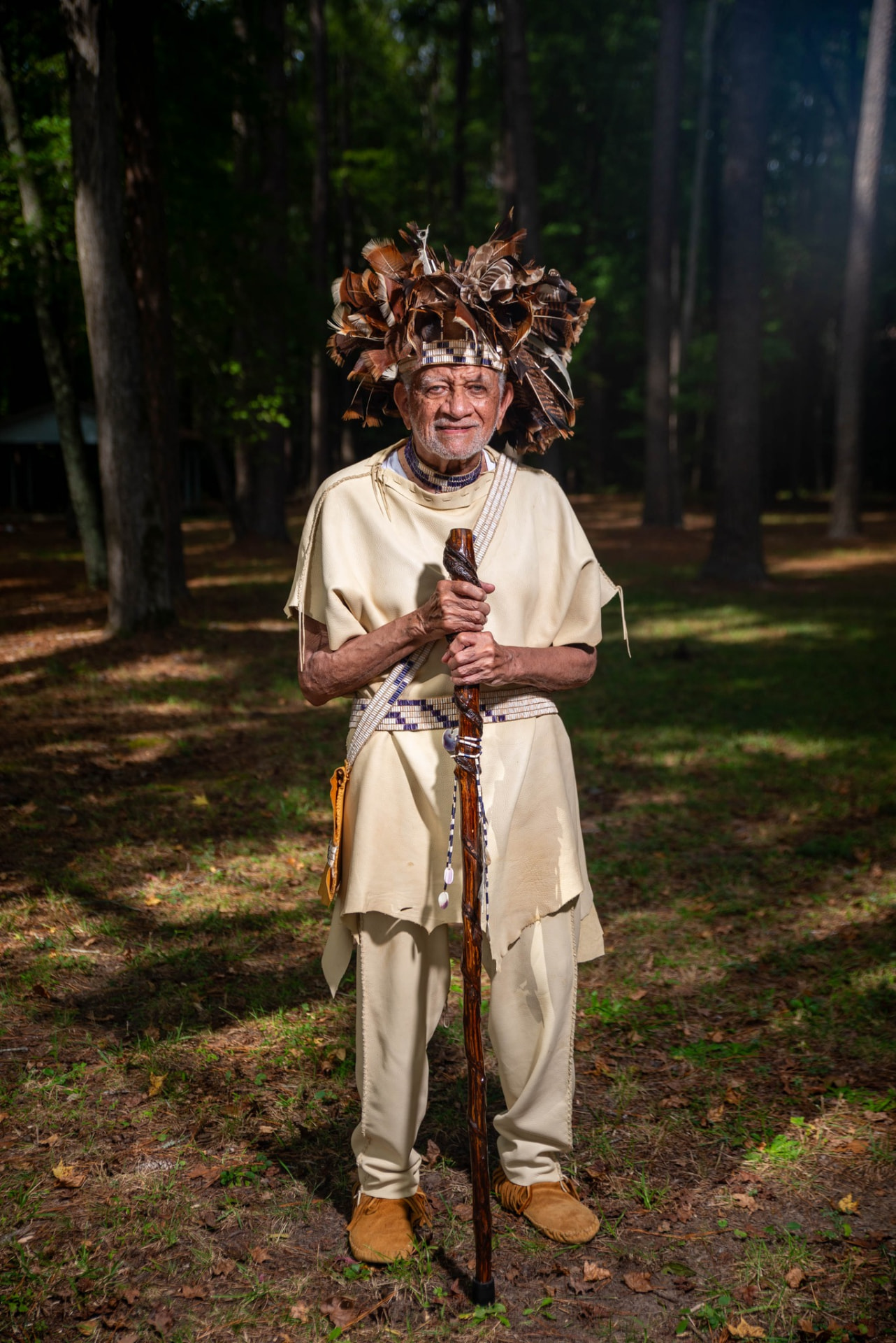 An elder in buckskin clothing and a feathered headdress standing in the woods holding a carved wooden staff