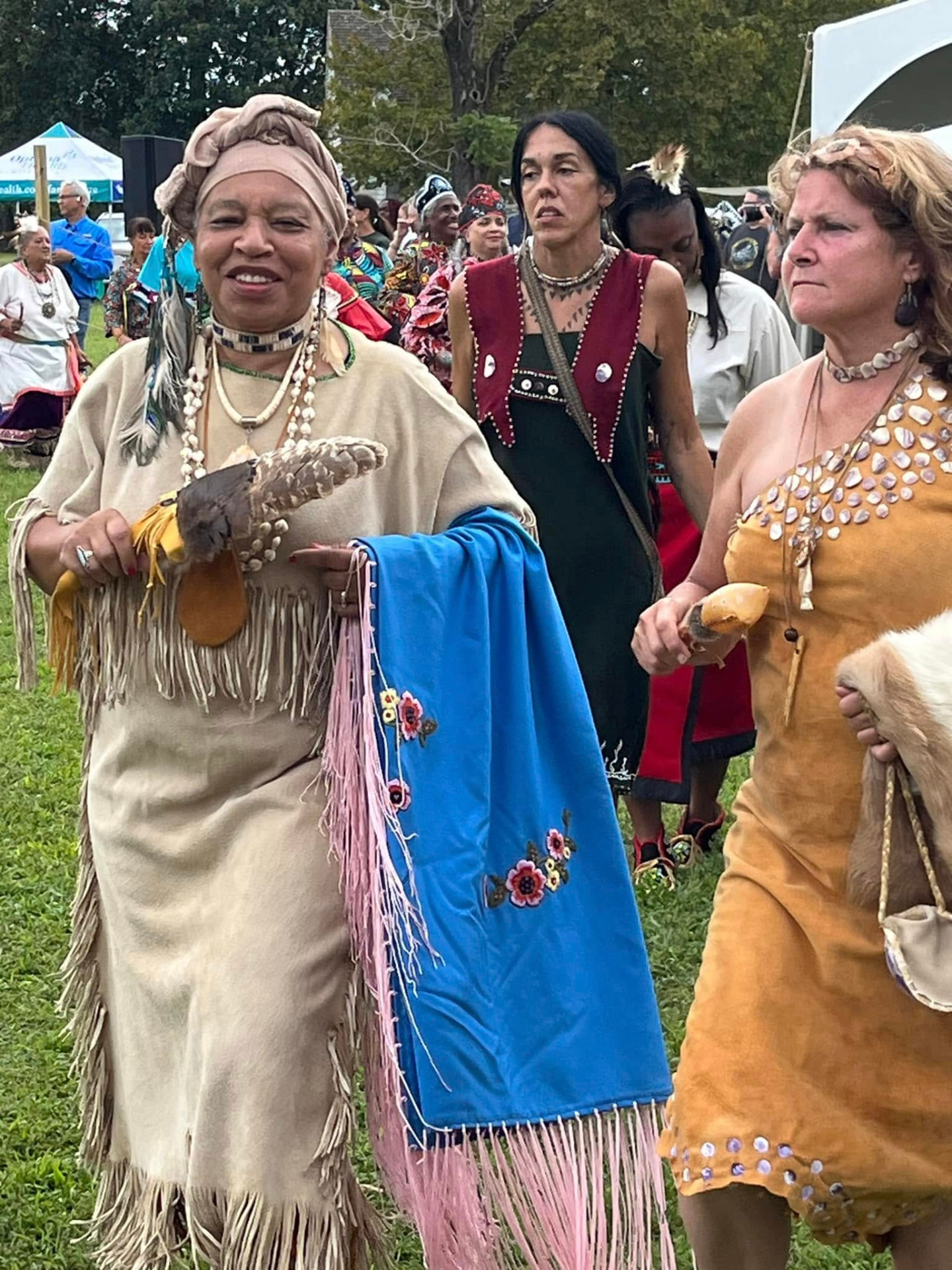 A smiling woman in a tan fringed buckskin dress and head wrap carries a blue shawl with pink fringe and a feathered fan, leading a procession across a grassy field