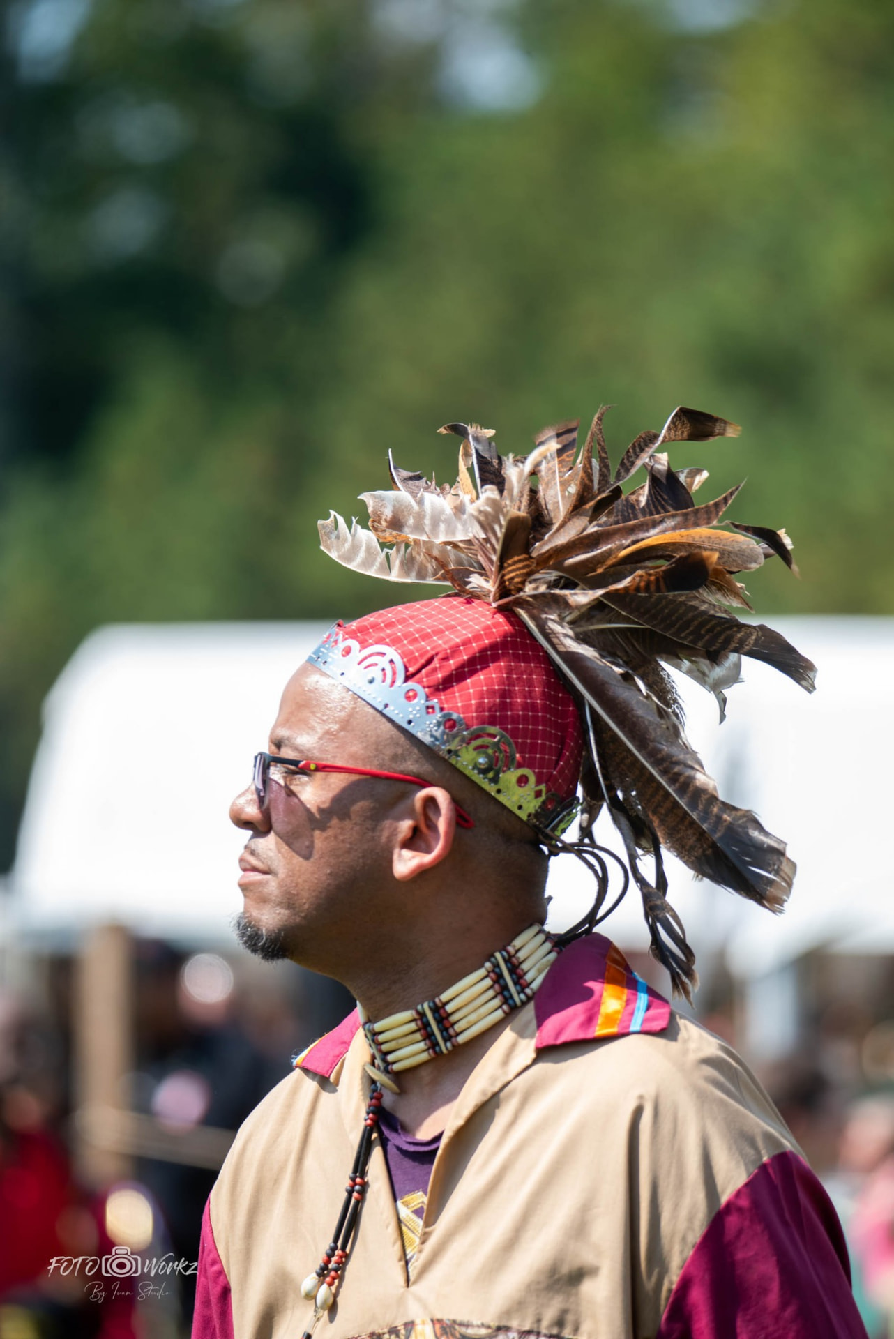 A man in profile wearing a red patterned headwrap with a fan of brown and white feathers, a tan shirt with maroon shoulder panels, and a bone choker necklace