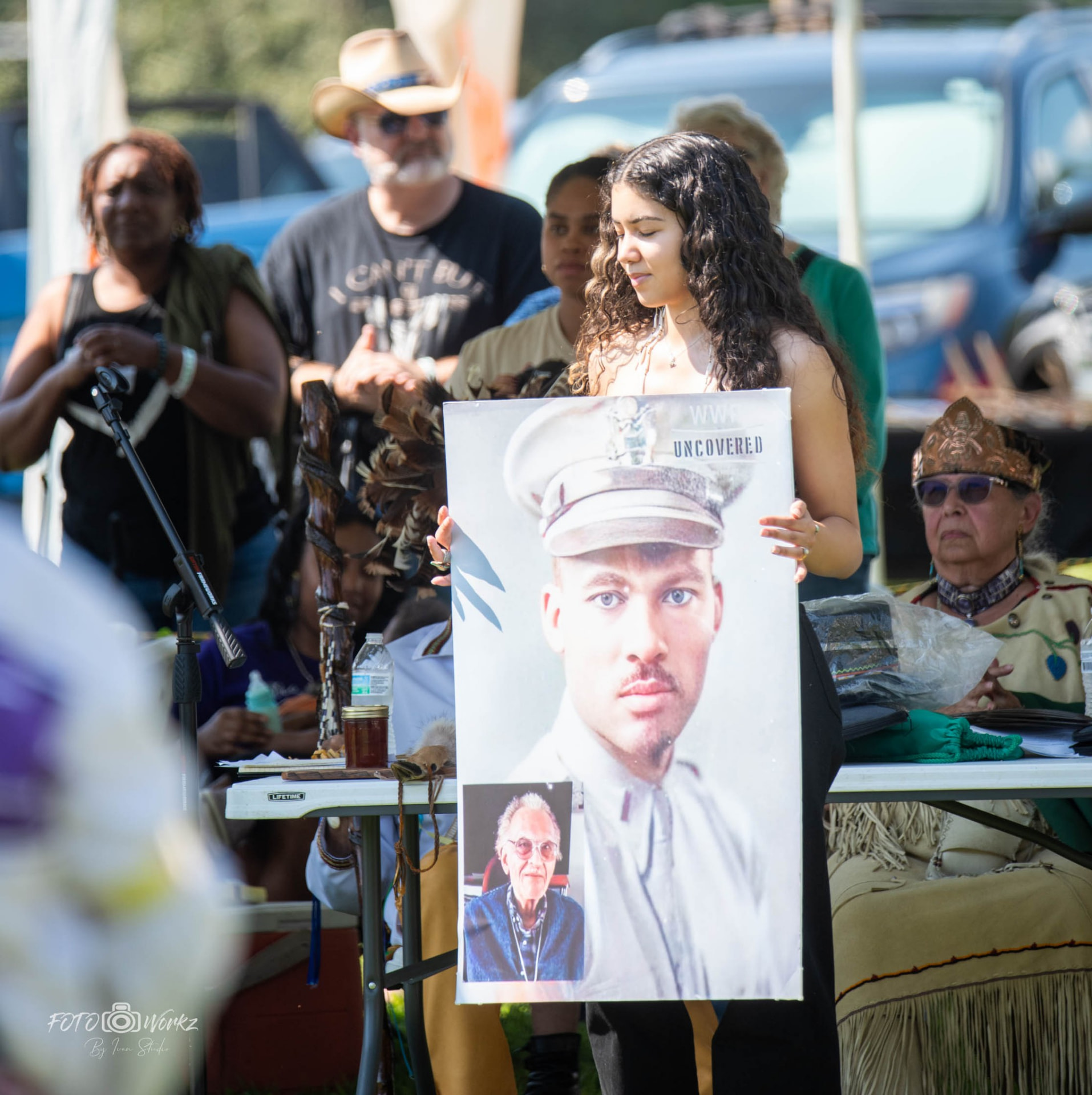 A young woman holding a poster titled WWII Uncovered showing a man in military uniform, at an outdoor tribal event