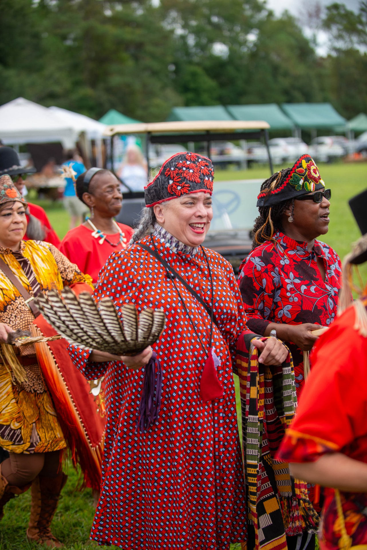Several women in red patterned dresses and beaded headpieces walk in a line across a grassy field, the woman in front holding a large striped feather fan