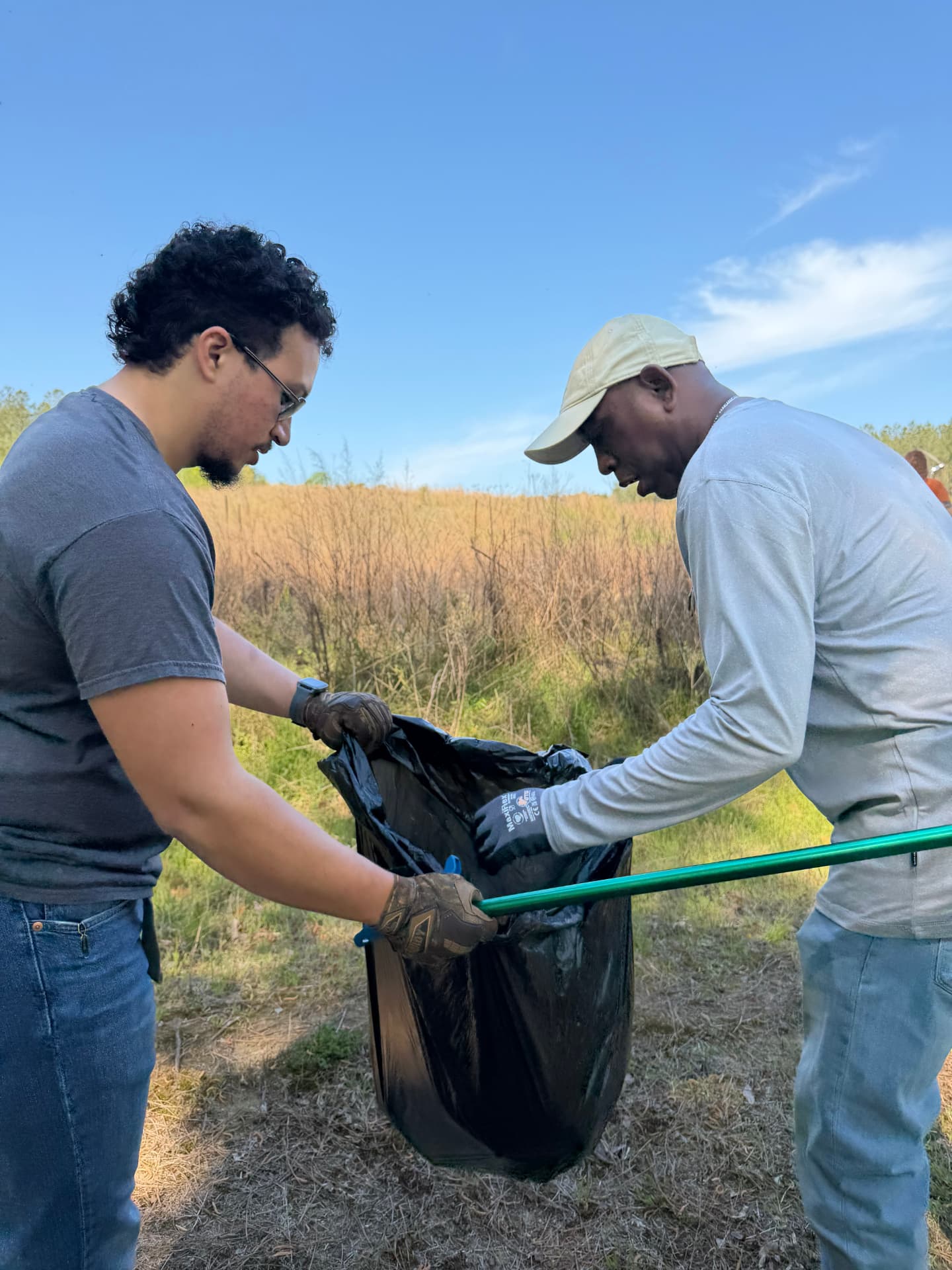 Two volunteers filling a black trash bag beside a grassy field