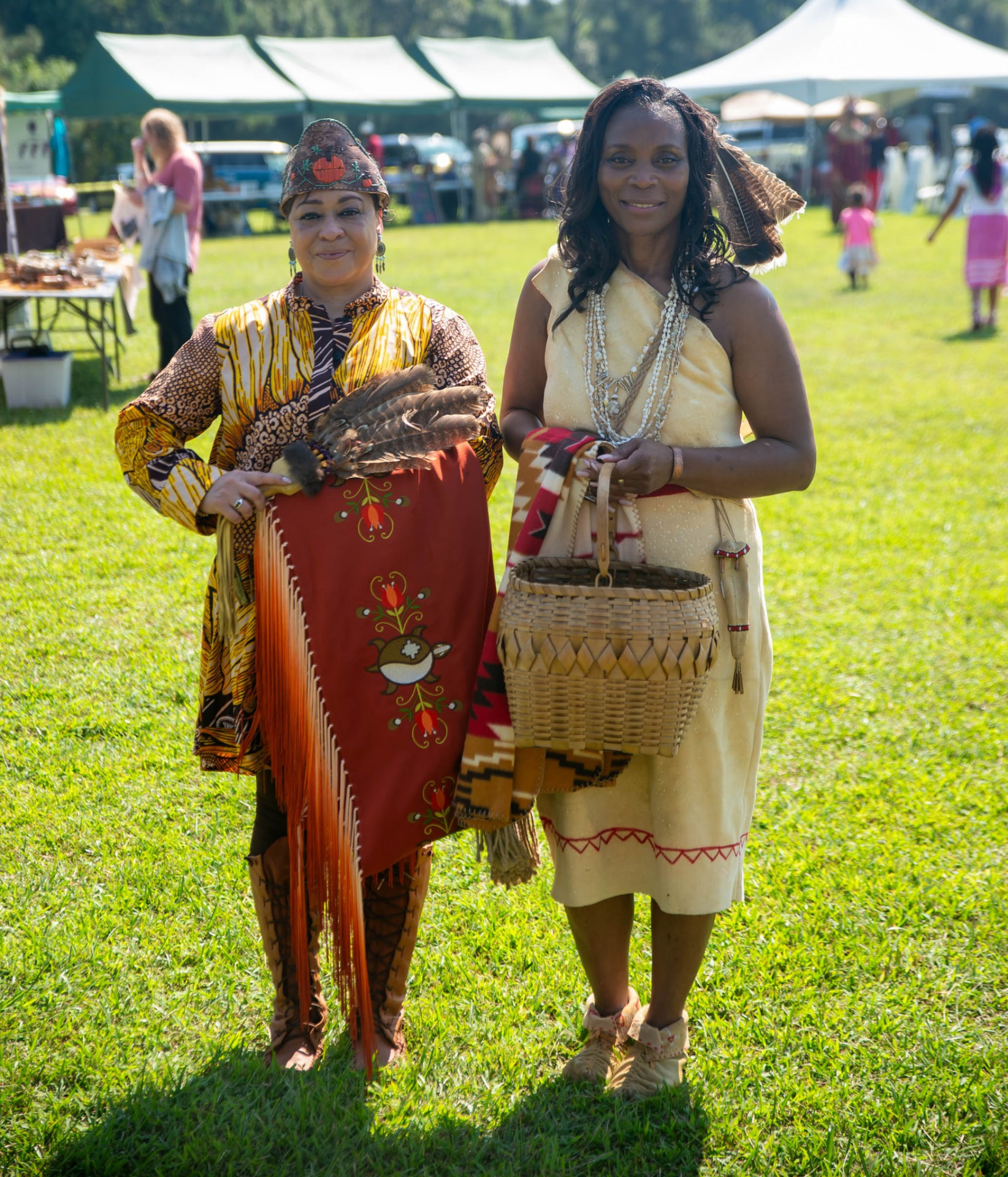 Two women standing in a field, one holding an orange fringed shawl and the other holding a woven basket and a folded blanket