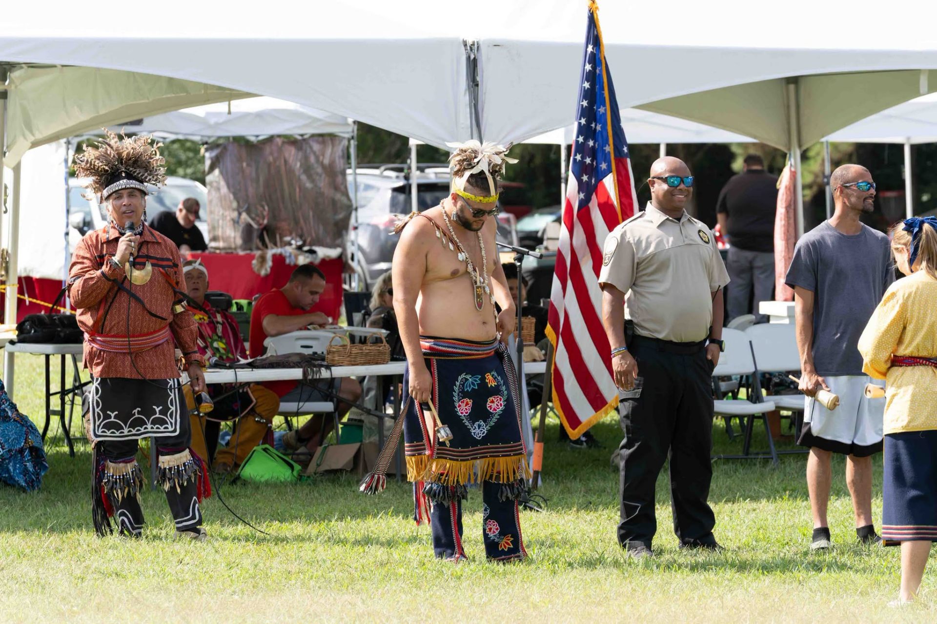 An elder singing into the microphone while a younger man stands before the American flag during a song at the powwow