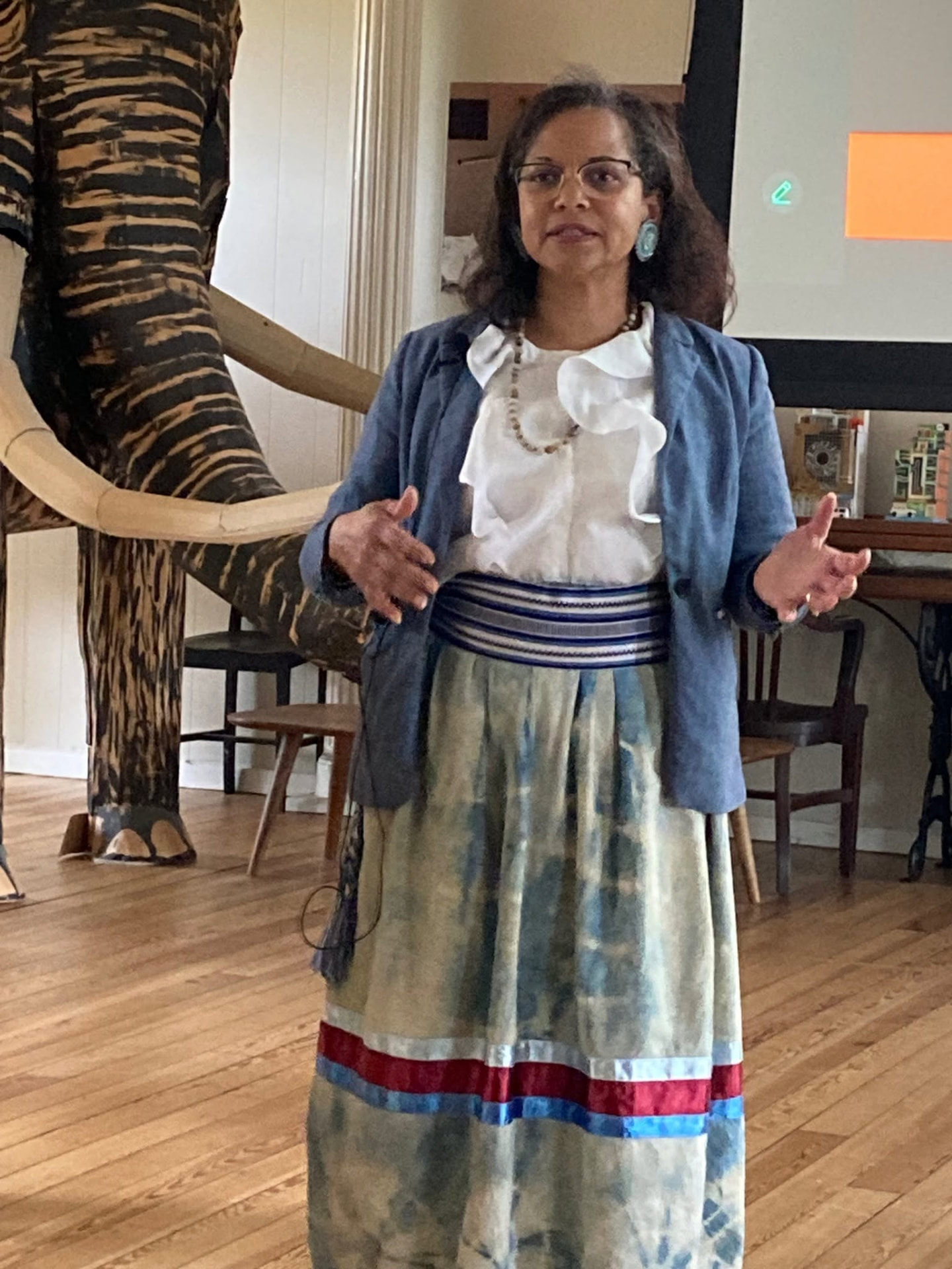 A woman in a blue jacket and a long skirt with red and blue ribbons gestures as she speaks at the Nottoway language presentation