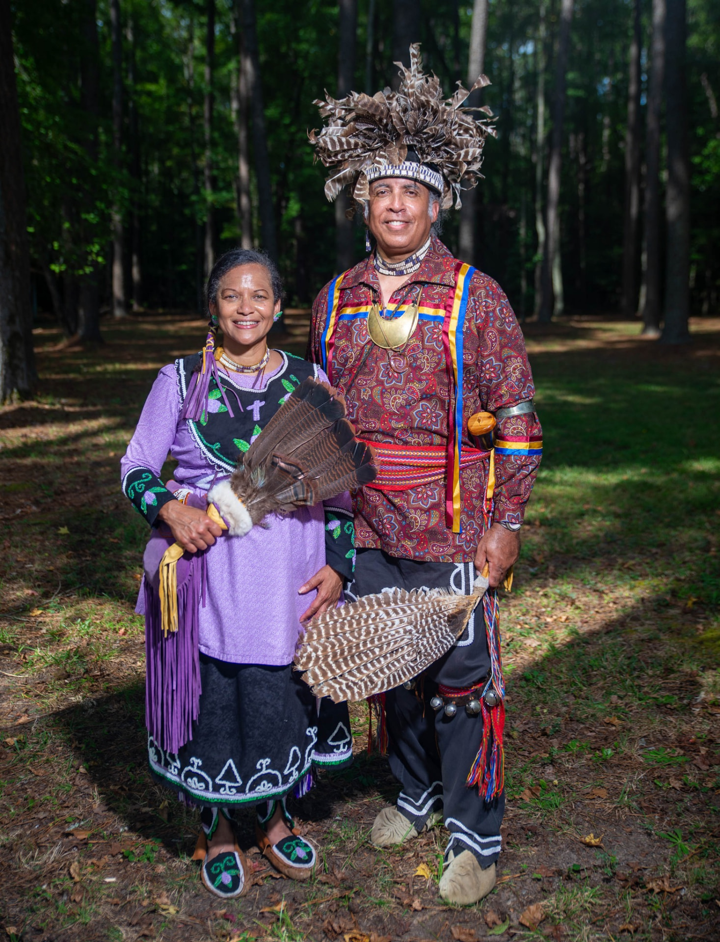 A woman in a lavender fringed tunic holding a feather fan and a man in a large feathered headdress standing together in the woods