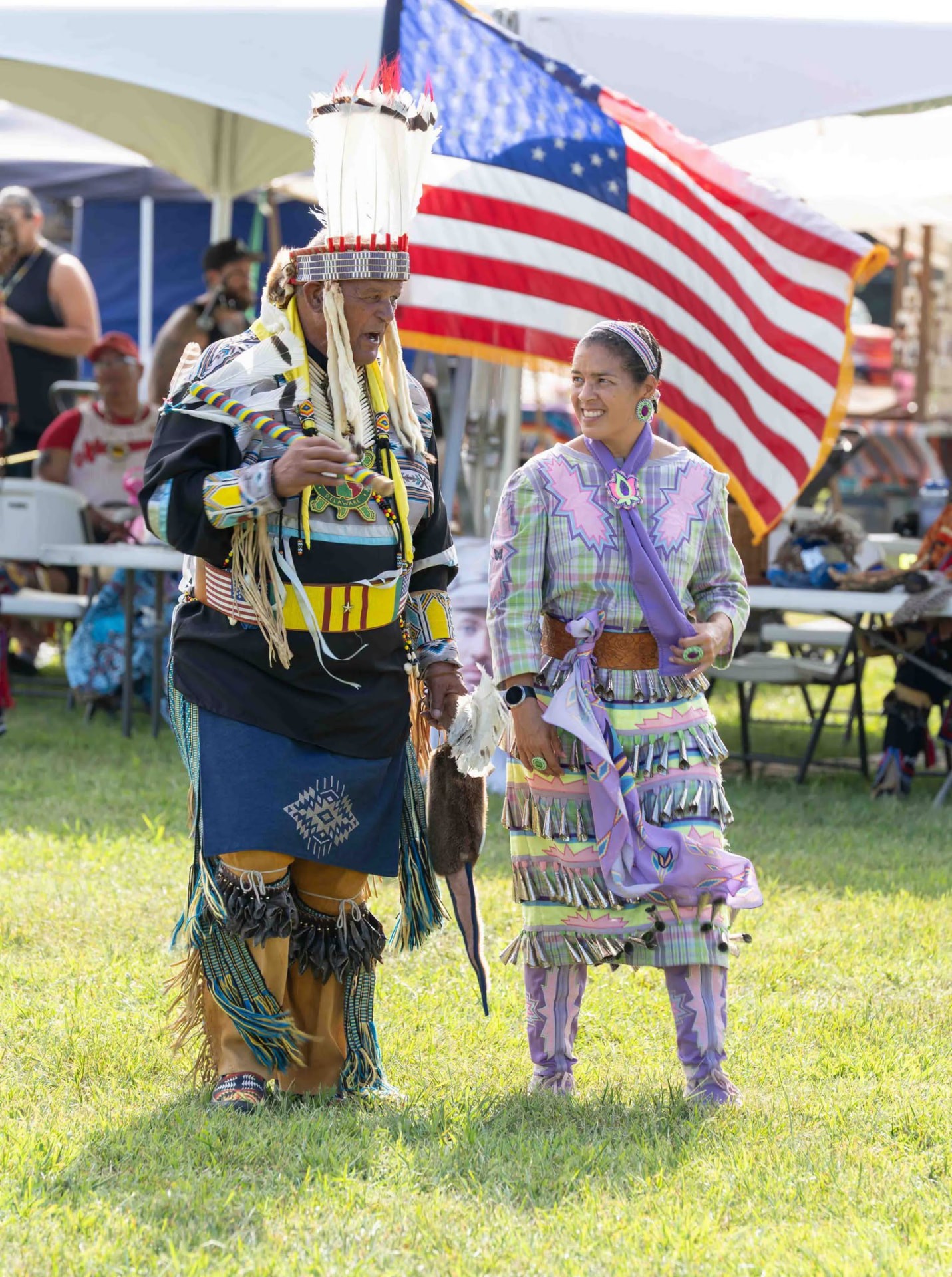 An elder in a feathered headdress talking with a jingle-dress dancer on the powwow grounds, an American flag behind them