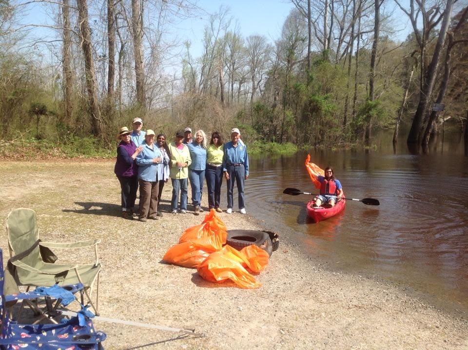 Volunteers standing together on the gravel shore at Cary's Bridge Landing after the annual cleanup, with filled orange trash bags and a discarded tire in front of them and a kayaker in the river holding up a bag of litter