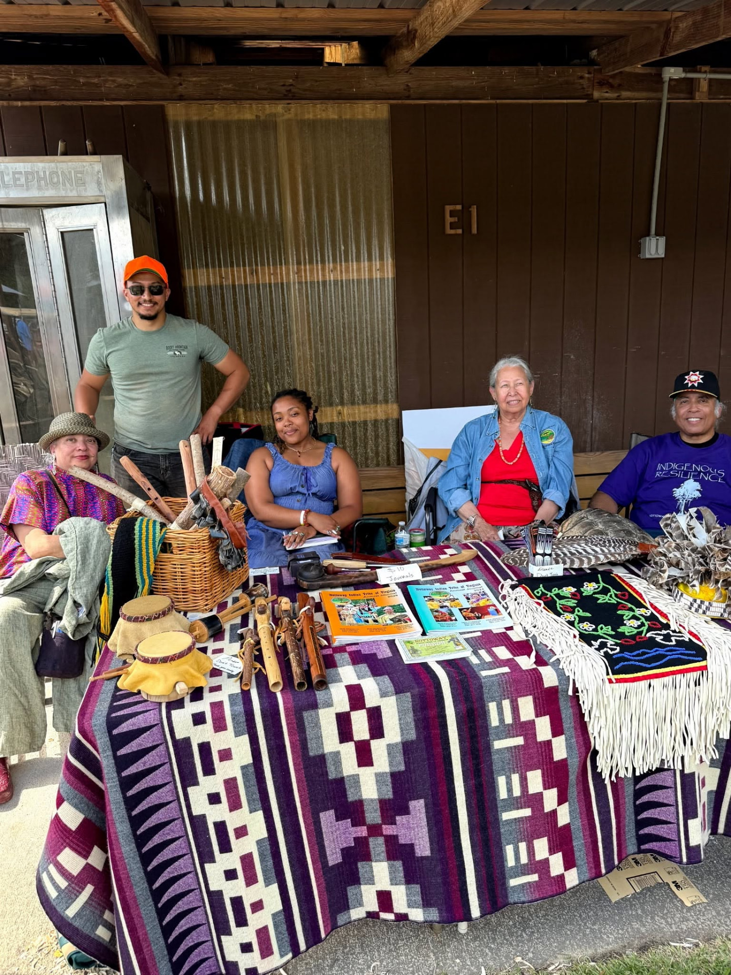 Tribe members and friends seated behind the Nottoway Indian Tribe of Virginia booth, with journals, drums, flutes, and a woven display on the table