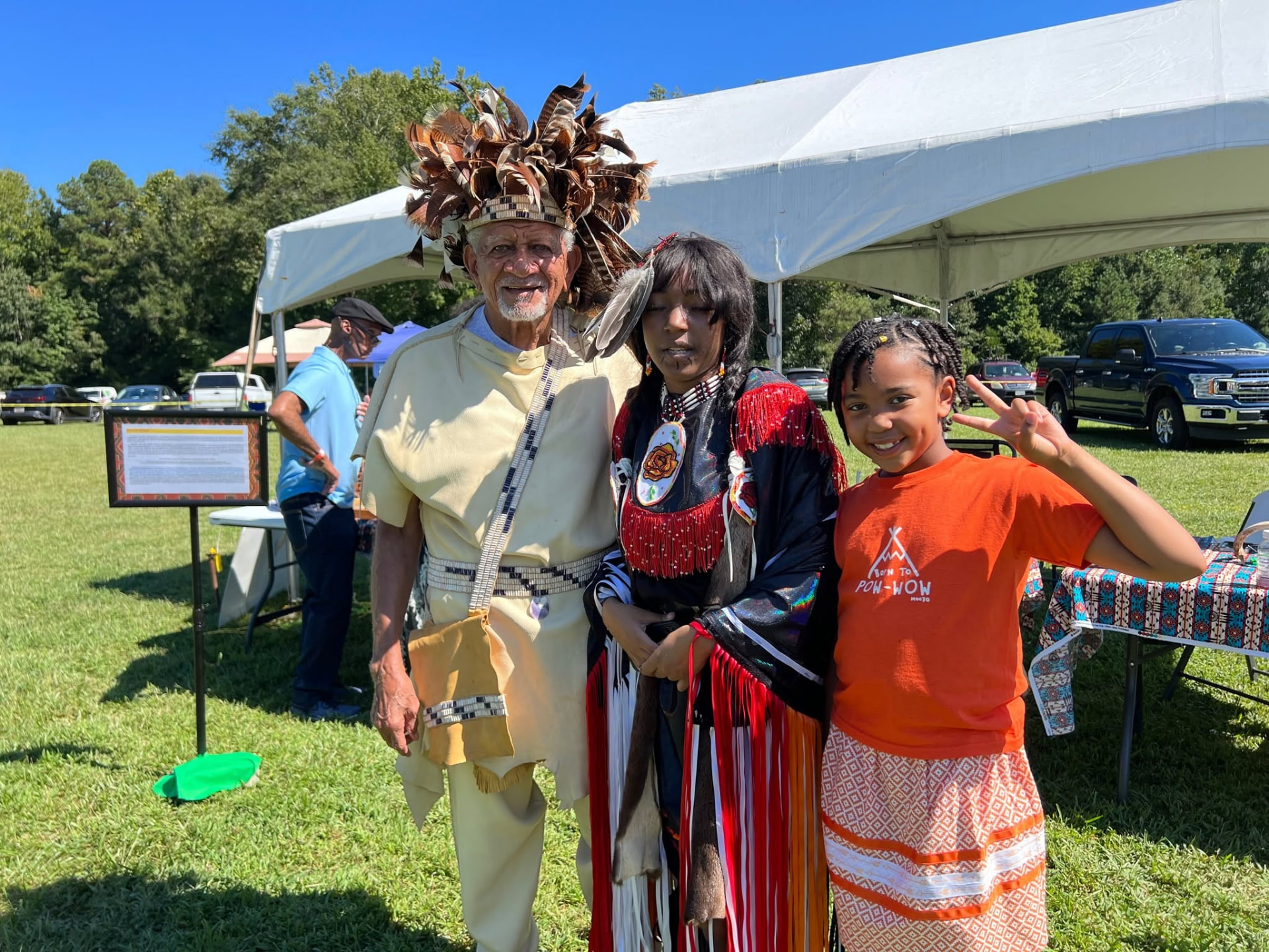 A man in a feathered headdress, a woman in fringed regalia, and a young girl in a Born to Pow-Wow shirt posing together outdoors