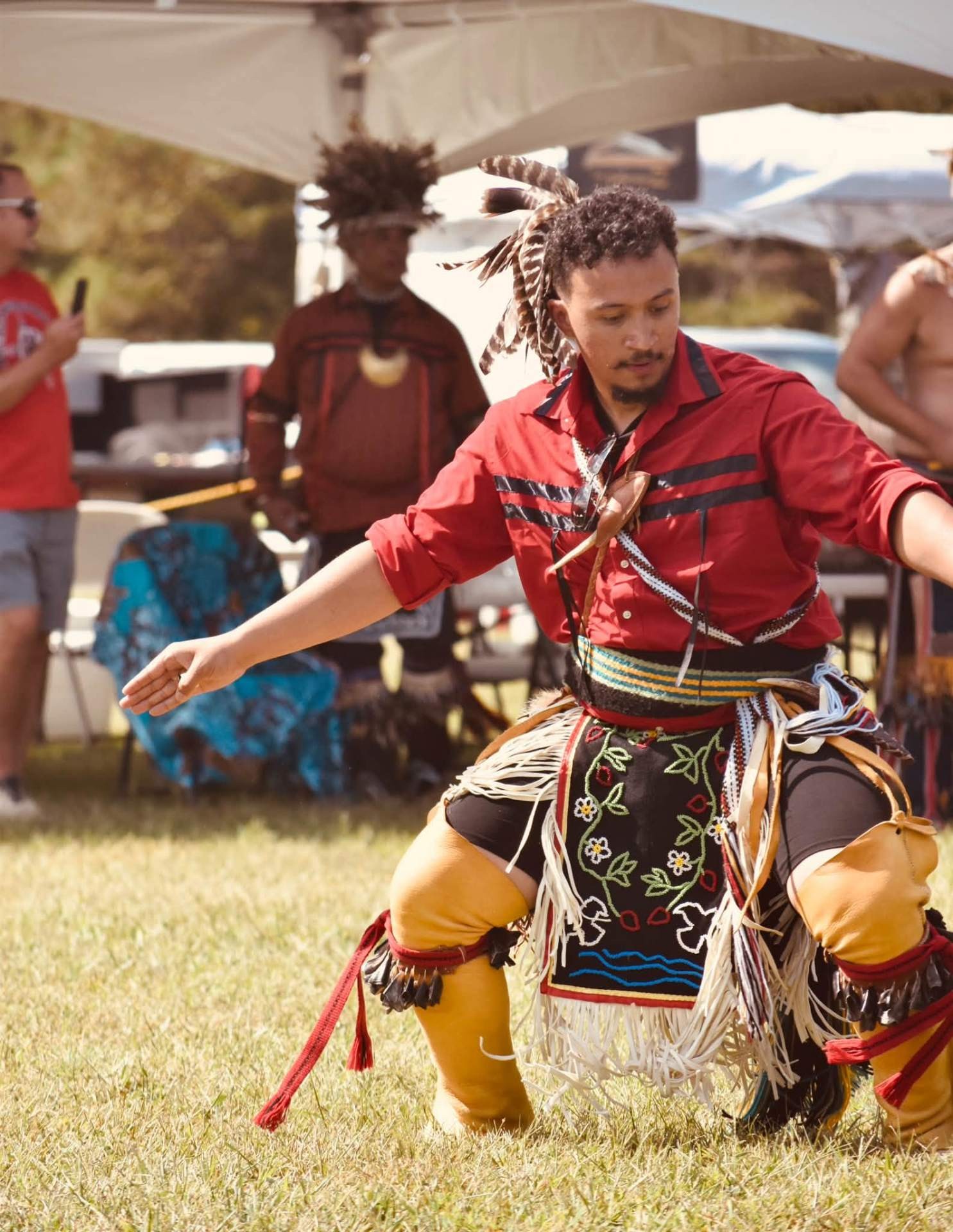 A dancer in a red shirt and beaded apron dancing low to the ground