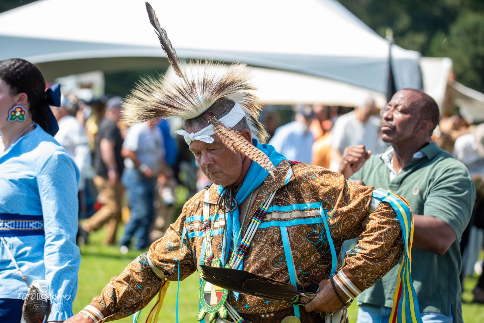 A man wearing a white headwrap with a feather crest and a beaded chest medallion reading Nanticoke and Delaware, holding a feather fan at an outdoor gathering