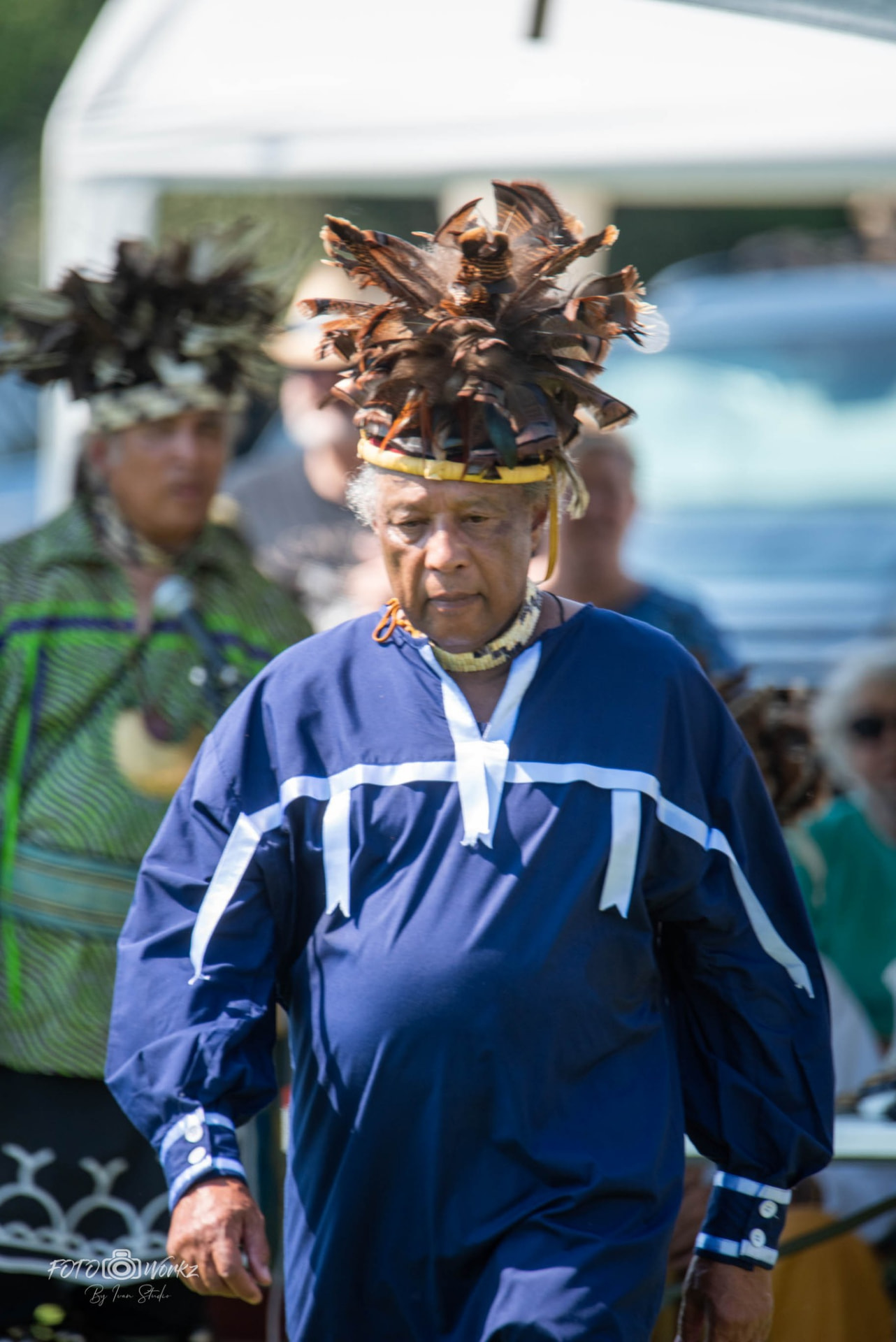 A man in a navy tunic with white trim and a feathered headdress walking forward at an outdoor powwow