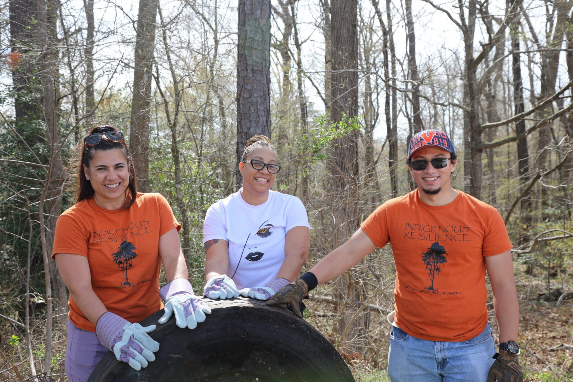 Three smiling volunteers leaning on a salvaged tire in the woods