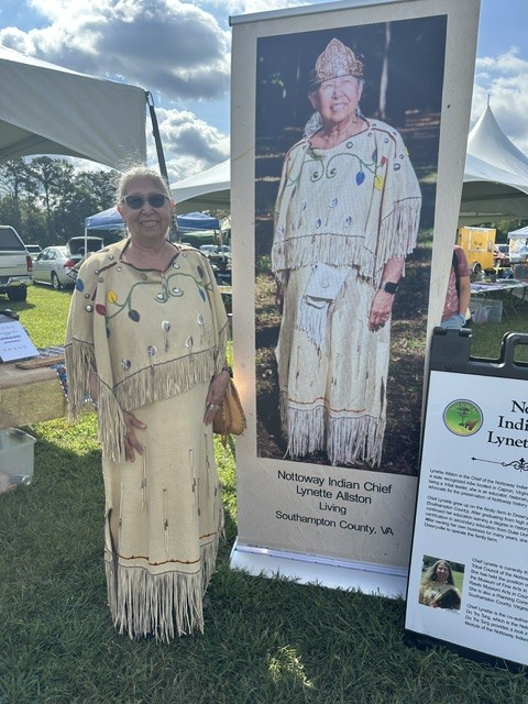 Chief Lynette Allston standing beside her display banner in her beaded regalia