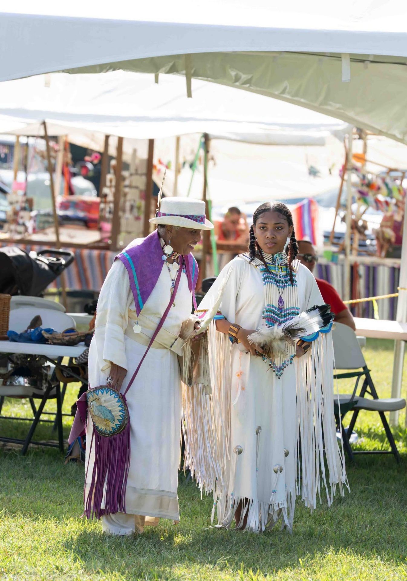 An elder in a white hat and purple shawl walking beside a young woman in white fringed regalia