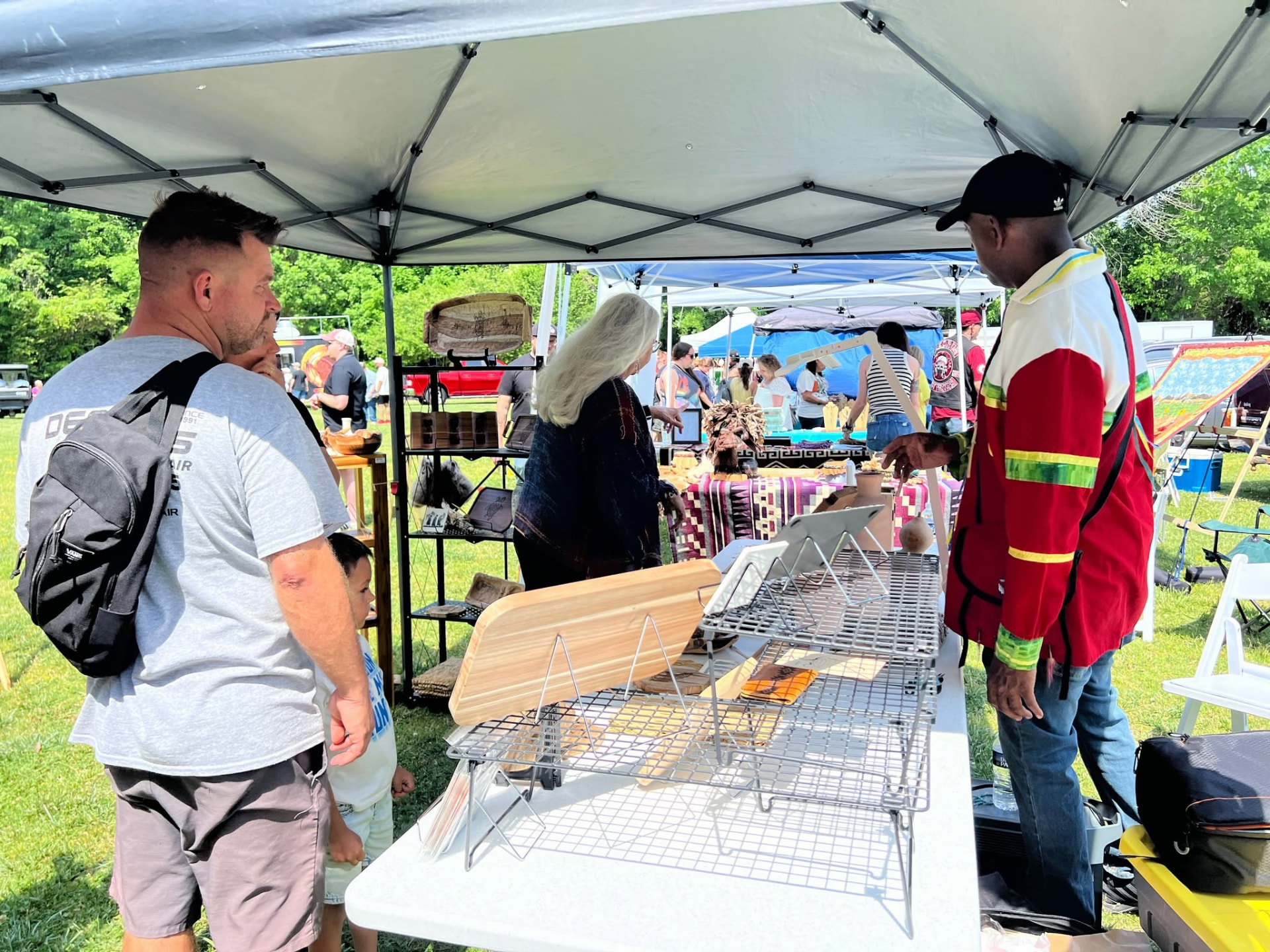 Visitors browse a craft booth under a white canopy at the Jamestown Indigenous Arts Festival, with woven baskets on a shelving unit and wooden boards on the table