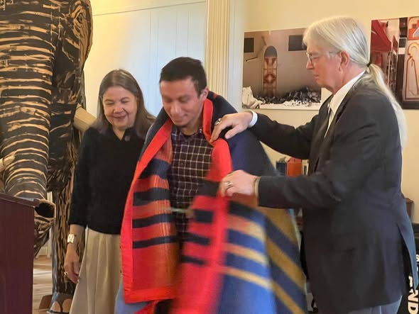 An honor blanket with red, blue, and gold stripes is placed around a man's shoulders during the Native American Student Graduation Program, as another person looks on