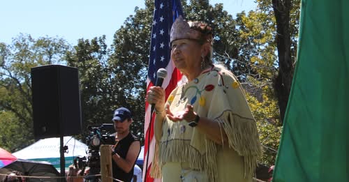 A woman in fringed regalia speaking into a microphone with an American flag behind her at the 2025 powwow